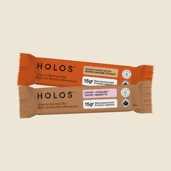 Two HOLOS organic nutrition bars in wrapper packaging, one orange and one brown, displayed on a solid light beige background.